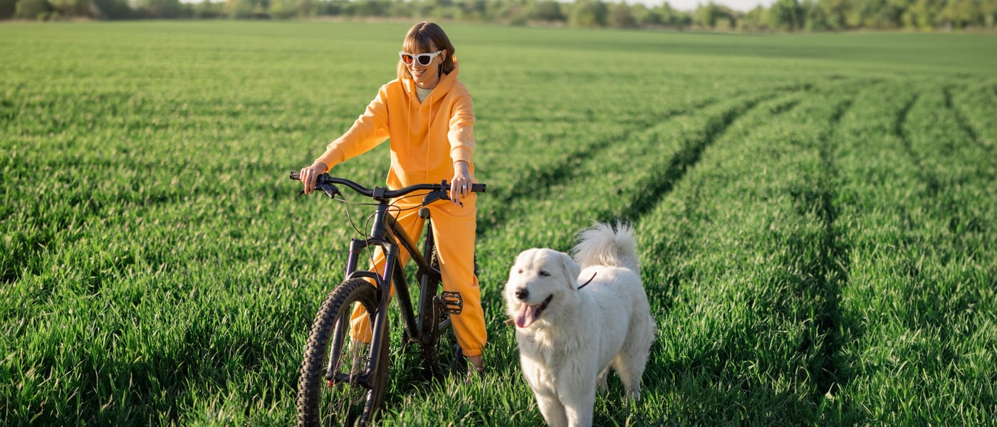 a lady in sunglasses and an orange tracksuit sitting on a bike in a grassy field with a retriever next to her
