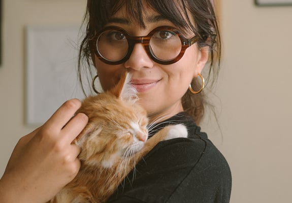 A woman with dark hair and glasses sitting on a couch holding an orange kitten.