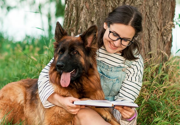 A girl reading a book against the tree with her German Shepherd in her arms