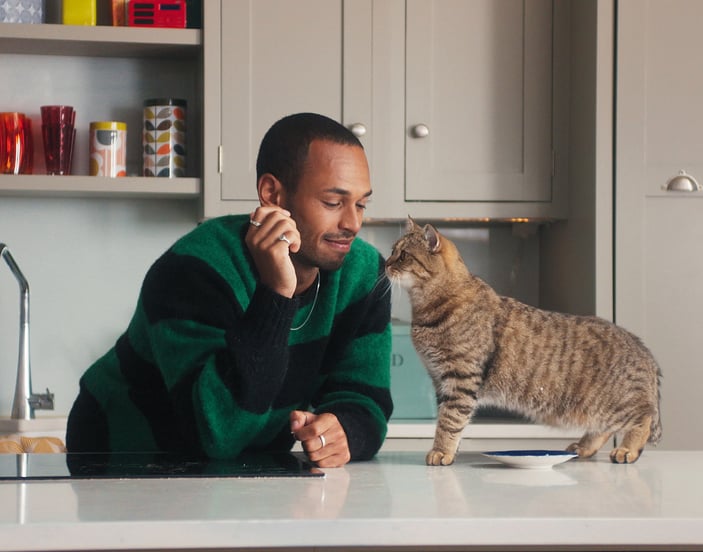 A man leaning on his kitchen counter and playing with his tabby cat.