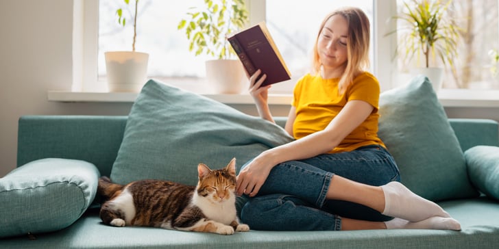 A woman sitting on a sofa reading a book next to her cat.