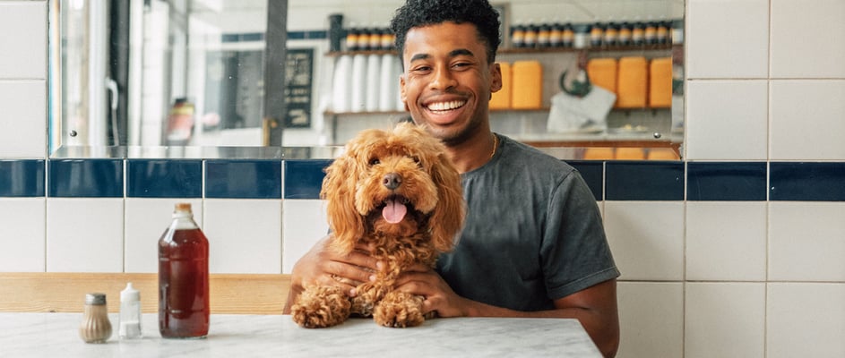 A man sits with his dog in a pie shop, they're both facing the camera with happy expressions.