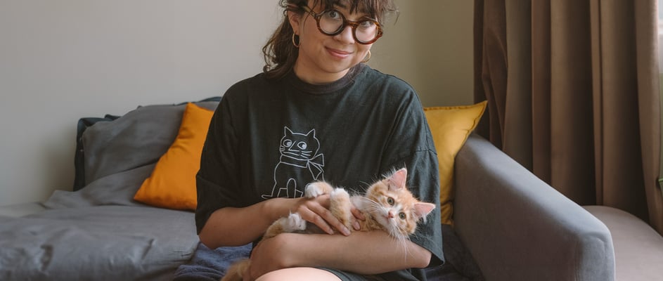 A woman with dark hair and glasses sitting on a couch holding an orange kitten.